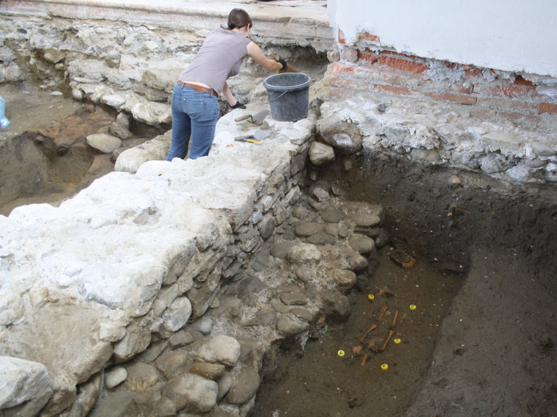 Angath - Mitarbeiterin beim Putzen der gotischen Mauer, im Vordergrund ältere überbaute Mauerteile und ein Kindergrab im Friedhof